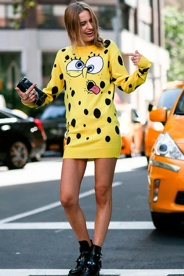 Person wearing a yellow sweater dress with a cartoon face design, posing on a city street with taxis in the background.