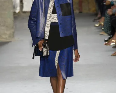 Model walking the runway in a blue patterned jacket and skirt with black accents, carrying a small handbag.