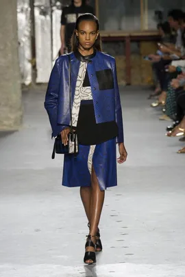 Model walking the runway in a blue patterned jacket and skirt with black accents, carrying a small handbag.