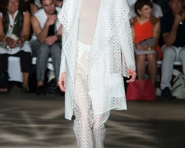 Model in white lace outfit walks runway at fashion show, audience in background.