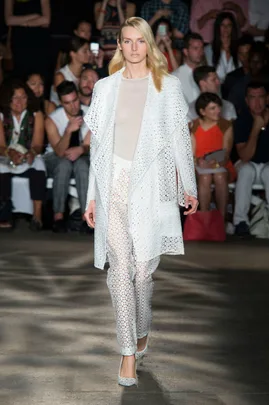 Model in white lace outfit walks runway at fashion show, audience in background.