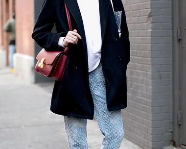 Person wearing a black coat, gray beanie, patterned jeans, blue sneakers, and a red bag standing in an urban setting.