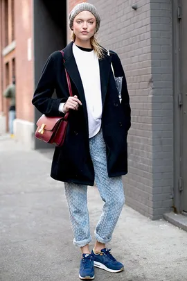 Person wearing a black coat, gray beanie, patterned jeans, blue sneakers, and a red bag standing in an urban setting.