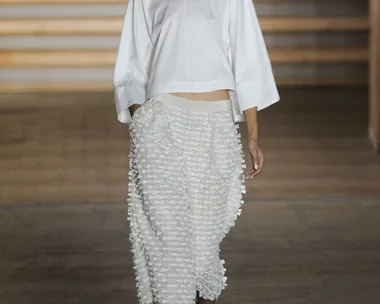 Model wearing a white top and textured skirt walks down a runway with wooden panel background.