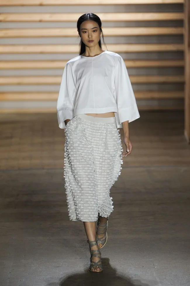 Model wearing a white top and textured skirt walks down a runway with wooden panel background.