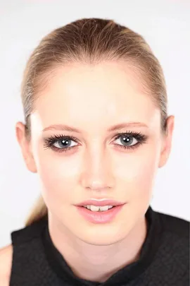 Close-up of a woman with blue eyes, light makeup, and a black sleeveless top, gazing directly at the camera, with hair tied back.