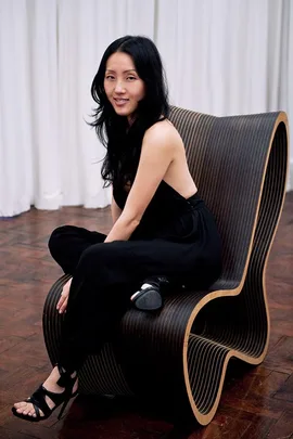 A woman in a black outfit sits on a modern, wavy wooden chair, posing and smiling with a white curtain in the background.