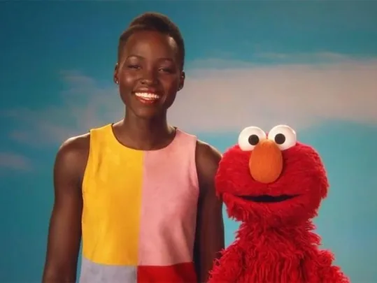 Lupita Nyong'o in a colorful dress standing next to Elmo from Sesame Street, both smiling against a blue sky background.