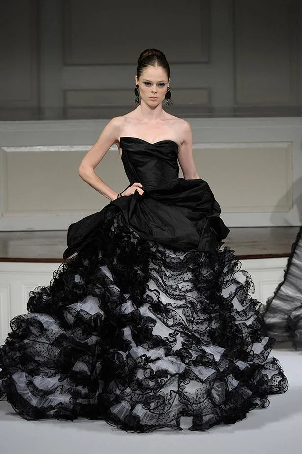 A model walks the runway in an elegant, strapless black Oscar de la Renta gown with ruffled layers and lace details.