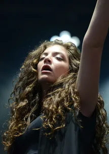 Singer Lorde with curly hair and eyes closed, performing on stage with an arm raised.