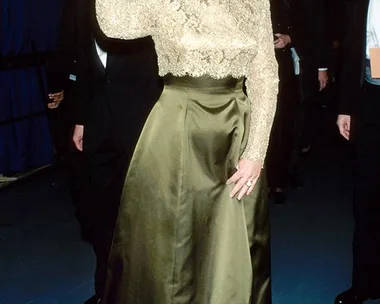 A woman waves, wearing a gold lace top and olive skirt at an event, surrounded by people in formal attire.