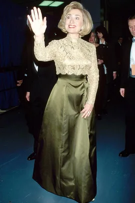 A woman waves, wearing a gold lace top and olive skirt at an event, surrounded by people in formal attire.