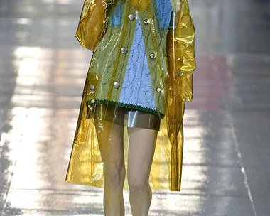 A model walks the runway in a blue and green knit top under a translucent yellow raincoat during a fashion show.