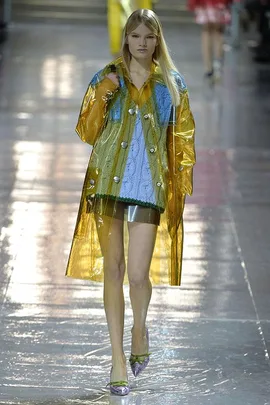 A model walks the runway in a blue and green knit top under a translucent yellow raincoat during a fashion show.