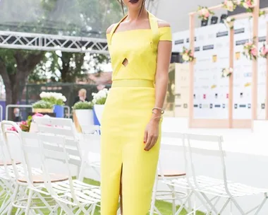 A woman in a yellow dress and black heels poses outdoors at the 2014 Caulfield Cup, with white chairs in the background.