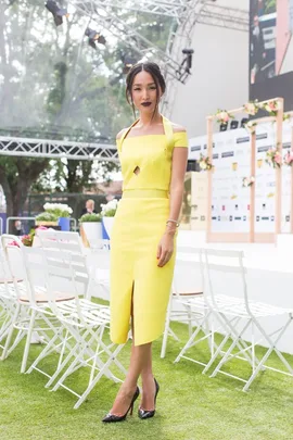 A woman in a yellow dress and black heels poses outdoors at the 2014 Caulfield Cup, with white chairs in the background.