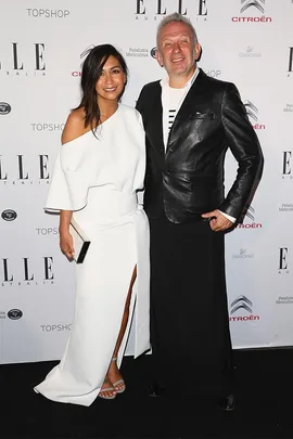 Jean-Paul Gaultier and a woman smiling at an event, he wears a leather jacket and she wears a white dress.