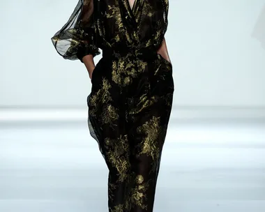 Model in a black and gold asymmetrical dress walks confidently down the runway with hands in pockets.