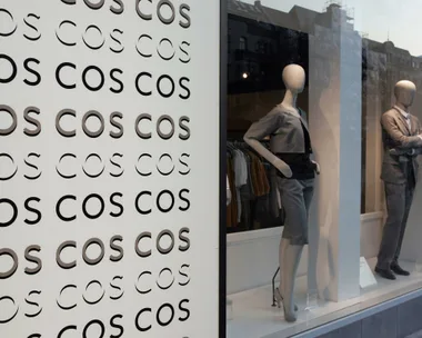 "Storefront of COS with mannequins in fashionable business attire and repeated 'COS' logo on display."