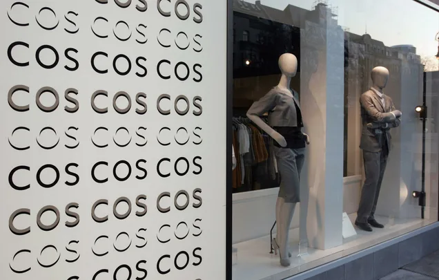 "Storefront of COS with mannequins in fashionable business attire and repeated 'COS' logo on display."