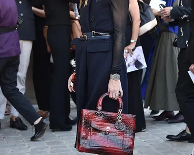 Olivia Palermo at a fashion event wearing a dark sleeveless dress, red handbag, and sunglasses, with a crowd in the background.