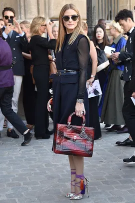 Olivia Palermo at a fashion event wearing a dark sleeveless dress, red handbag, and sunglasses, with a crowd in the background.