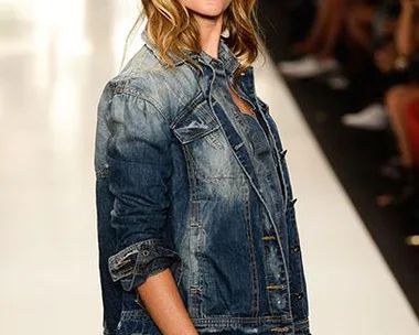 Woman in denim jacket and jeans, hands in pockets, walks confidently on a fashion runway with blurred audience in background.