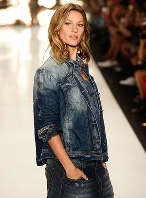 Woman in denim jacket and jeans, hands in pockets, walks confidently on a fashion runway with blurred audience in background.