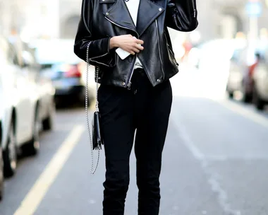 A woman in a black leather jacket and boots stands on a city street, smiling and making a peace sign.