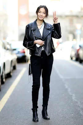 A woman in a black leather jacket and boots stands on a city street, smiling and making a peace sign.