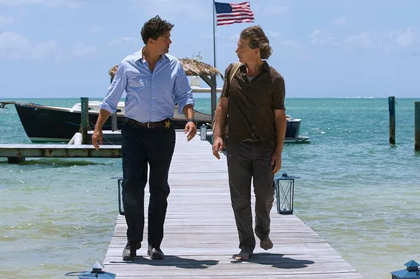 Two men walk on a dock over clear blue water, American flag flying in background.