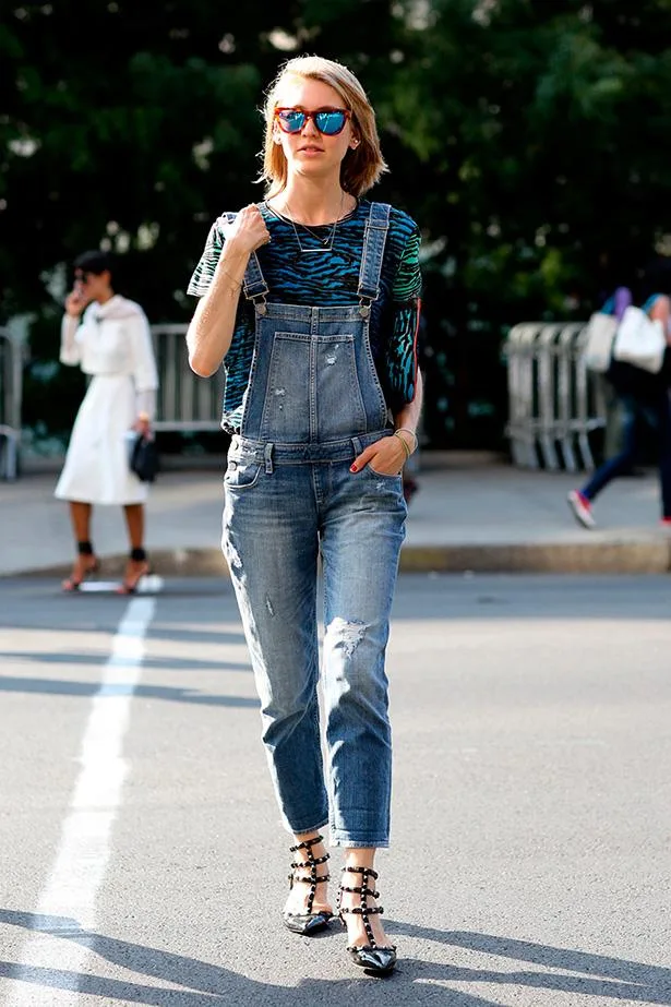 A person in denim overalls and colorful sunglasses walks on a sunny street.