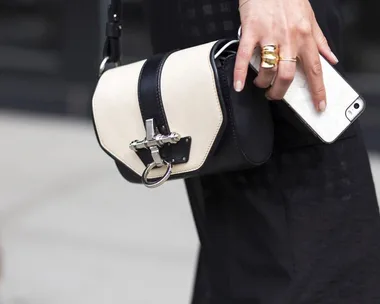 A person holding a small black and white crossbody bag and a smartphone, wearing gold rings.