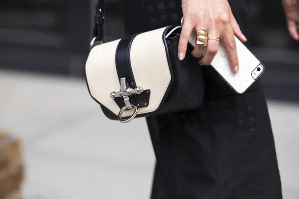 A person holding a small black and white crossbody bag and a smartphone, wearing gold rings.