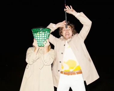 Two people in beige coats; one has a green basket on their head, the other holds a golf club and appears surprised.