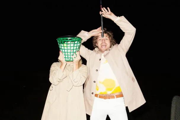 Two people in beige coats; one has a green basket on their head, the other holds a golf club and appears surprised.