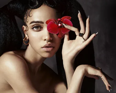 A person with intricate hair holds a red flower near their eye, wearing hoop earrings and a nose ring, against a dark background.