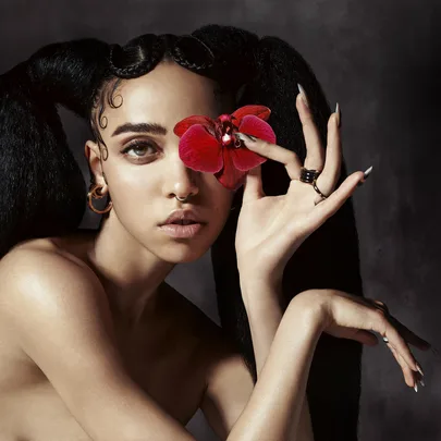 A person with intricate hair holds a red flower near their eye, wearing hoop earrings and a nose ring, against a dark background.