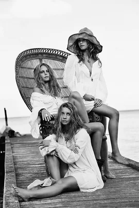 Three women in white dresses pose on a wooden pier, with one sitting on a wicker chair and another wearing a large hat.