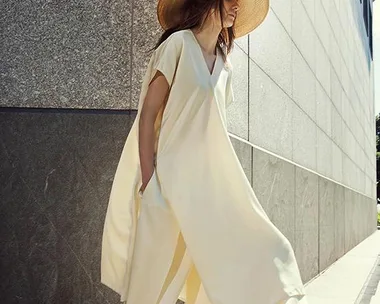 A woman in a flowing cream dress and wide-brimmed straw hat walks along a modern building.