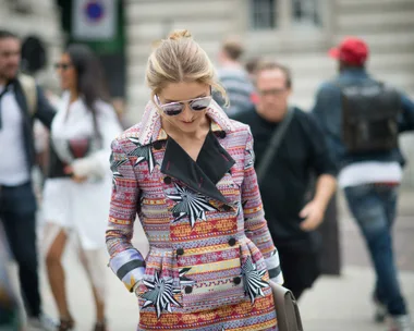 Fashionably dressed woman in a vibrant patterned coat walking on the street, wearing sunglasses.