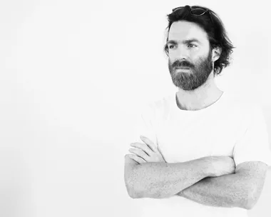 Man with beard in white T-shirt and sunglasses on head, arms crossed, looking away in black and white photo.