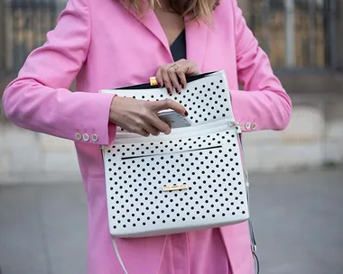 Woman in pink blazer holding a white, polka-dotted purse and placing an object into it.