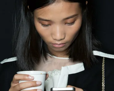 Young woman with black hair looks down at her phone while holding a white coffee cup, wearing a black and white outfit.