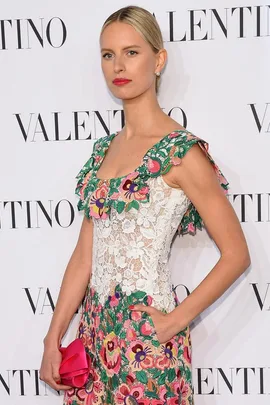 A woman in a floral lace dress poses with one hand in a pocket, at a Valentino event, holding a pink clutch.