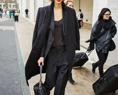 Woman in a stylish black outfit walking confidently outdoors, holding a black bag, with people in the background.