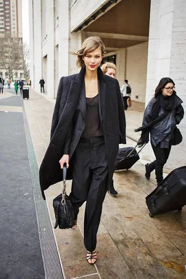 Woman in a stylish black outfit walking confidently outdoors, holding a black bag, with people in the background.