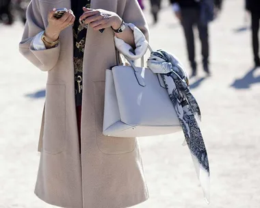 Woman in beige coat holding a phone and large white handbag with a patterned scarf; blurred background with people walking.