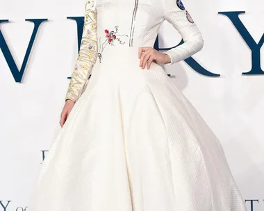 A woman in a detailed white dress with floral embroidery, posing on a blue carpet at a film premiere event.