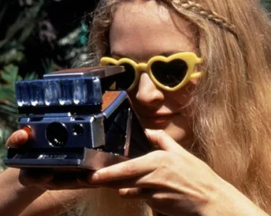 Blonde woman in heart-shaped sunglasses taking photo with vintage Polaroid camera outdoors.